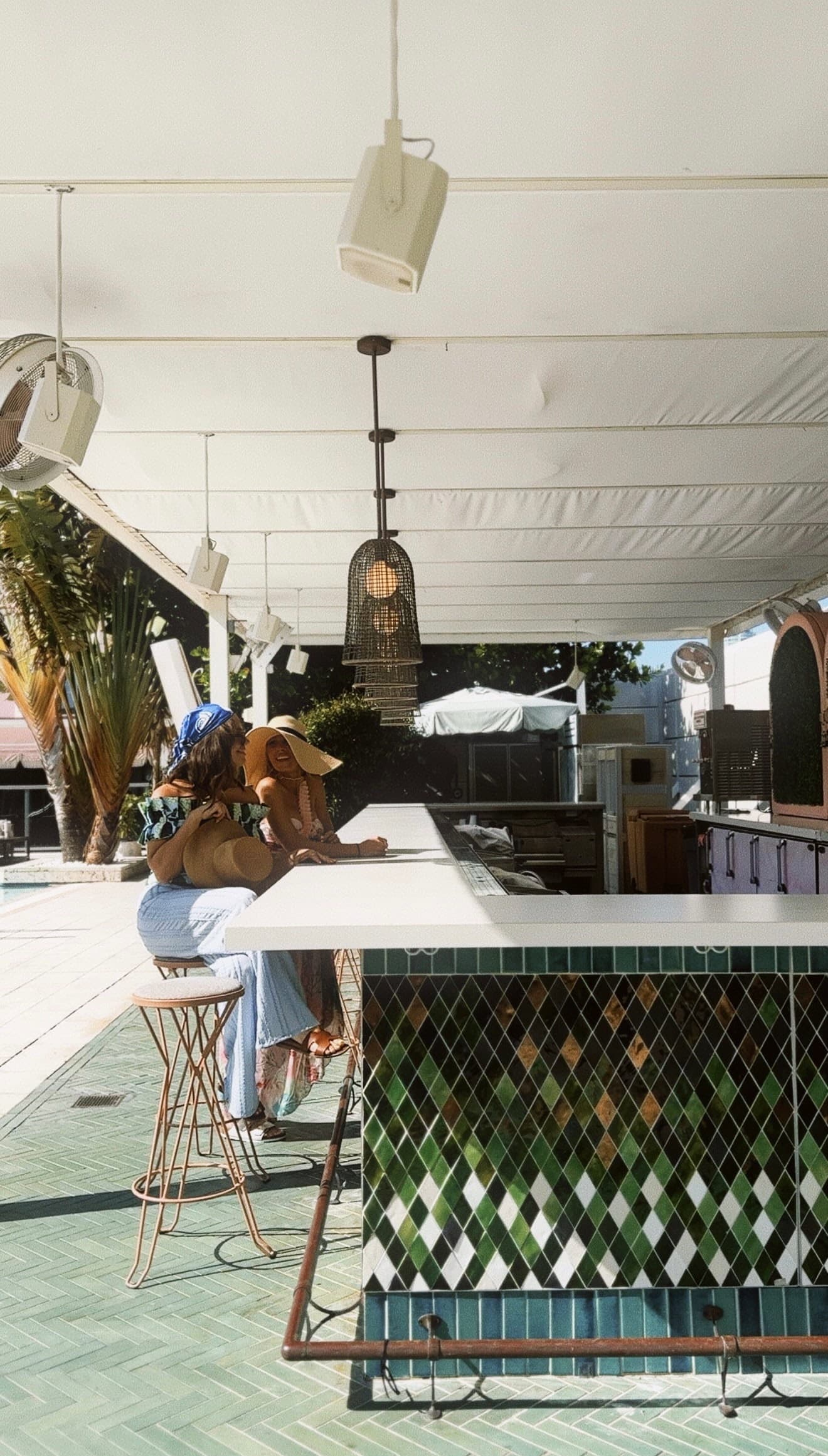 Two friends laughing together at a sunlit poolside bar with green tilework and palm trees